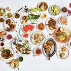 Vielfältige asiatische Gerichte: gegrilltes Fleisch, Nudeln, Reis, Melonenscheiben, bunte Teller und bunte Blumen im Hintergrund.