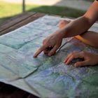 Text links oben: "KOMPASS". Zwei Hände zeigen gemeinsam auf eine ausgebreitete topografische Karte auf einem Holztisch.