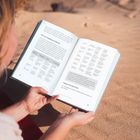 Ein aufgeschlagenes Buch mit Tabellen und Texten über "Alternative Antiplattitüden". Hände halten das Buch auf Sand.
