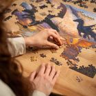 Eine Hand setzt ein Puzzle mit Drachenmotiv zusammen. Man sieht viele unvollständige Teile auf einem Holztisch.