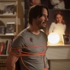 Ein Mann mit langem Haar und Bart ist mit einem orangefarbenen Kabel gefesselt. Im Hintergrund stehen Bücher und Fotos.