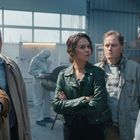 Mehrere Personen diskutieren in einem Labor. Ein Mann trägt Schutzanzug und Maske, im Hintergrund arbeiten Forscher.