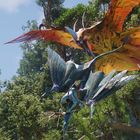 Eine blaue und orange Kreatur fliegen über dichten Dschungel; Detailreiche Flügel und Bäume im Hintergrund.