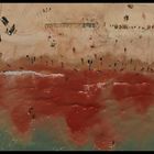 Am Strand bewegen sich Menschen und Fahrzeuge. Das Wasser erscheint rot, als ob es mit Farbe oder Blut vermischt ist.