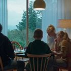 Vier Personen sitzen an einem Tisch in einem Raum, der gemütlich beleuchtet ist. Ein großes Fenster zeigt Bäume draußen.