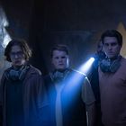 Personen in einem dunklen Raum, tragen Atemschutzmasken, blaues Licht im Hintergrund. Wand mit blauem Graffiti.