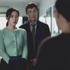 Eine Frau in einem grünen Cardigan und ein Mann im Anzug schauen ernst zu einer dritten Person.