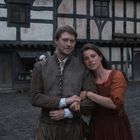 Ein Paar in historischer Kleidung steht vor einem Fachwerkhaus auf einem Hof mit einem Hund und weiteren Personen im Hintergrund.