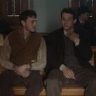 Zwei Männer in altertümlicher Kleidung sitzen auf einer Holzbank, umgeben von anderen Personen in einem Warteraum.