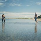 Zwei Männer in Badehosen stehen im flachen Wasser an einem sonnigen Strand mit blauem Himmel.