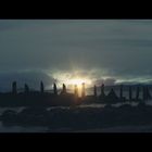 Kein Text sichtbar. Sonnenuntergang hinter alten Holzpfählen im Wasser, dunkle Wolken und ruhige Meeresoberfläche.