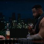 Eine Frau und ein Mann sitzen in einer Bar, vor ihnen eine Flasche Wein, Nachtstadt im Hintergrund.