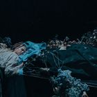 Eine Frau in historischer Kleidung liegt an einem Sarg mit Blumen. Die Stimmung ist dunkel und melancholisch.