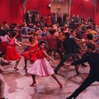 Viele tanzende Paare in bunter Kleidung in einem Raum mit roten Wänden, lebhaftes Ambiente.