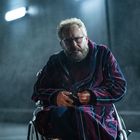 Ein Mann mit Brille und Bart sitzt in einem Rollstuhl, trägt einen gestreiften Bademantel, es ist dunkel und regnerisch.