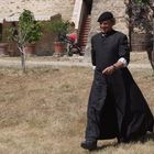 Ein Mann in schwarzer Priesterkleidung und Barrett geht über trockenes Gras, hinter ihm ein altes Steinhaus mit Pflanzen.