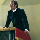 Ein Mann in einem Priestergewand steht lächelnd am Rednerpult, mit rotem Tuch bedeckt, schlichte Umgebung.