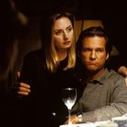 Eine Frau umarmt einen Mann; sie sitzen an einem Tisch mit Weinglas und Tassen. Beide wirken ernst.