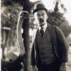 Ein Mann mit Bowlerhut und Schnurrbart steht komisch schauend neben einem Strauß in einem Park.