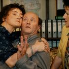 Drei Personen in einem Büro. Eine Frau umarmt einen Mann, der überrascht wirkt; eine zweite Frau beobachtet sie. Im Hintergrund Aktenordner.