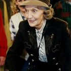 Eine ältere Frau in einem eleganten, bestickten Sakko und Hut lächelt. Menschen sind im Hintergrund zu sehen.