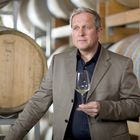 Ein Mann in einem braunen Anzug hält ein Weinglas vor Fässern in einem Weinkeller.