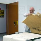 Ein Schild mit "Vorsicht Kunst" steht im offenen Türrahmen. Eine Person hält ein großes, braunes Papier mit Zeichnung.