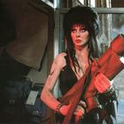 Eine Frau mit schwarzem Haar hält eine große rote Röhre in einem industriellen Umfeld. Rauch und Taschen im Hintergrund.