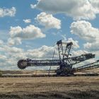 Großer Schaufelradbagger auf einem braunen Tagebaugelände, mit blauem Himmel und weißen Wolken.
