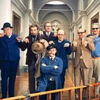 Person in blauer Jacke sitzt kniend, lächelt mit verschränkten Armen. Sechs Männer stehen hinter ihr in einer Innenaufnahme.