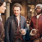 Drei Männer in historischen Kostümen mit Perücken, halten Weingläser, stehen in einem eleganten Raum mit Holzwänden.