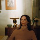 Eine Frau in einem warmen, beleuchteten Raum. Neben ihr eine Lampe und im Hintergrund ein gerahmtes Kunstwerk.