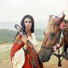 Frau in rotem Schal mit Blumenmuster hält eine Peitsche. Neben ihr ein Pferd, dessen Mähne mit Blumen geschmückt ist.