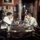 Zwei Männer spielen Schach in einem prunkvollen Raum, umgeben von antiken Möbeln und Dekoration.