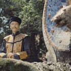 Ein Mann in traditioneller Kleidung steht neben einer kunstvollen Drachenstatue im Freien, umgeben von Bäumen.