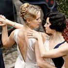 Zwei elegant gekleidete Frauen tanzen eng miteinander. Eine trägt ein helles, rückenfreies Kleid, die andere ein Kleid mit Perlenschmuck.