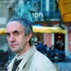 Nicht lesbares Schild im Hintergrund. Ein Mann mit grauem Haar und beigem Mantel steht draußen vor einem Café.