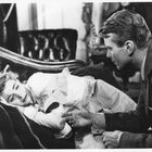 Ein Mann beugt sich besorgt zu einer Frau auf einem Sofa. Sie liegt mit geschlossenen Augen und hält eine Decke.