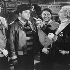 FSK-Kennzeichnung unten rechts. Vier Männer in verschiedenen Outfits, einer zeigt gestikulierend auf einen anderen.