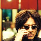 Ein Mann mit lockigem Haar und Sonnenbrille telefoniert, im Hintergrund rot-beigefarbene Unschärfe.