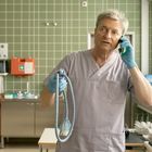 Ein älterer Mann in Krankenhausschürze telefoniert, hält ein Seil in der Hand, steht in einem Labor mit grün gekachelten Wänden.