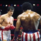 Zwei Boxer im Ring; einer in weiß-roten Shorts, der andere in rot-weiß-blauen. Ein Mann in rosa schaut zu.