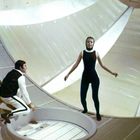 Zwei Personen in einem futuristischen, runden Raum; eine in Schwarz-Weiß-Kleidung, die andere in einem schwarzen Anzug.