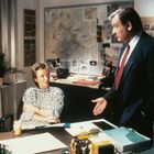 Zwei Personen in einem Büro. Links sitzt eine Frau mit kurzem Haar, rechts steht ein Mann im Anzug, eine Pinnwand im Hintergrund.