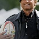 Ein Mann mit einer schwarzen Mütze und Lederjacke hält lächelnd einen Rugbyball. Er trägt eine Kette mit Kreuz.