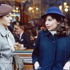 Zwei Frauen in Mänteln, eine trägt einen braunen, die andere einen blauen Hut. Sie stehen vor einem Café mit Menschen im Hintergrund.