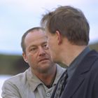 Zwei Männer sprechen miteinander im Freien. Einer schaut den anderen ernst an. Hintergrund ist unscharf.