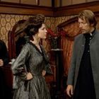 Ich sehe keine lesbaren Texte. Drei Personen in einem viktorianischen Raum. Eine Frau in einem eleganten Kleid spricht mit zwei Männern.