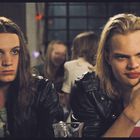 Zwei Personen mit langen Haaren und schwarzen Lederjacken sitzen an einem Tisch. Eine Person hält eine Zigarette.