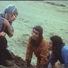 Drei Personen knien im Sand. Zwei tragen blaue Kleidung, eine braune. Eine zeigt gestikulierend nach vorne.